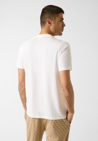 Maglietta bianca in cotone con scollo tondo, maniche corte e schiena liscia, indossata da una persona con pantaloni marroni a motivi.