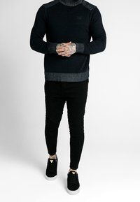 Suéter de cuello alto de punto oscuro con un diseño texturizado en el hombro, combinado con pantalones ajustados negros y zapatillas negras con suelas blancas.