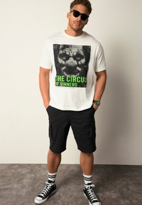 Camiseta blanca con un gráfico negro y verde de la cara de un payaso, combinada con pantalones cortos cargo negros, zapatillas altas negras y calcetas a rayas.