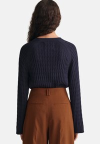 Frau mit langen, lockigen Haaren trägt einen marineblauen, grob gestrickten Pullover, der in hoch geschnittene, braune, weit geschnittene Hosen gesteckt ist und von hinten zu sehen ist.