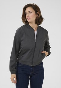 Graue Bomberjacke mit einem Frontreißverschluss, gerippten Bündchen und Saum. Glatte Stofftextur, kombiniert mit dunkelblauen Jeans.