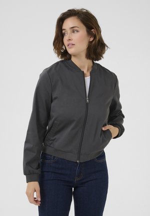 Graue Bomberjacke mit einem Frontreißverschluss, gerippten Bündchen und Saum. Glatte Stofftextur, kombiniert mit dunkelblauen Jeans.