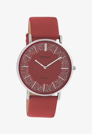 Montre ronde en argent avec bracelet en cuir rouge, cadran texturé rouge, index horaires argentés, et aiguilles des heures, minutes et secondes blanches.