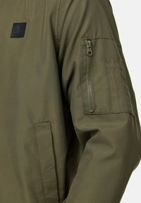 Manche de veste vert olive avec une poche zippée verticale et une poche à rabat rectangulaire sur le haut du bras, poche avant visible.