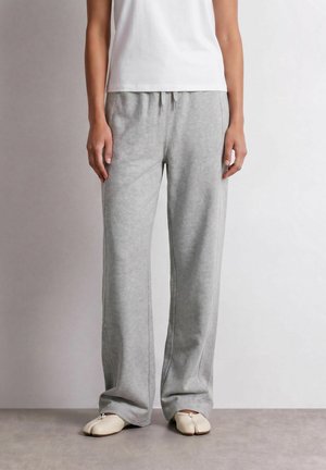 Person, die lockere graue Jogginghosen, ein weißes Oberteil und geteilte beige Schuhe trägt, steht auf einem glatten Boden vor einem schlichten Hintergrund.