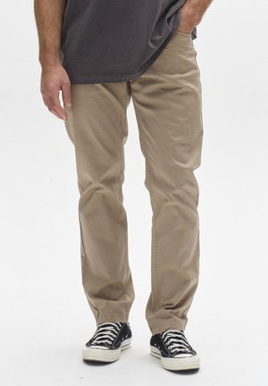 Mees, kes kannab beežid vabaajapüksid, mustad madala säärega tossud valgete paeltega ning tumehallist särki, ühe käega püksitaskus.