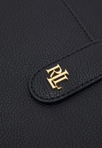 Svart läderaccessoar med en texturerad yta, med en detalj i guld med monogram. Designen inkluderar en rundad lockstängning.