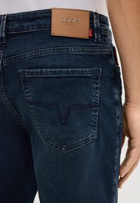 Jeans en denim bleu foncé avec un patch en cuir portant l'inscription "JOOP JEANS" sur la taille et une couture unique en forme de V sur la poche arrière.