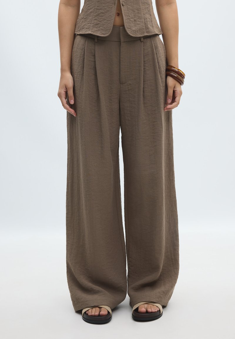 Pantalon marron à larges jambes avec plis, porté avec un gilet assorti et des sandales beiges à bout ouvert, accessoirisé avec plusieurs bracelets marron à un poignet.