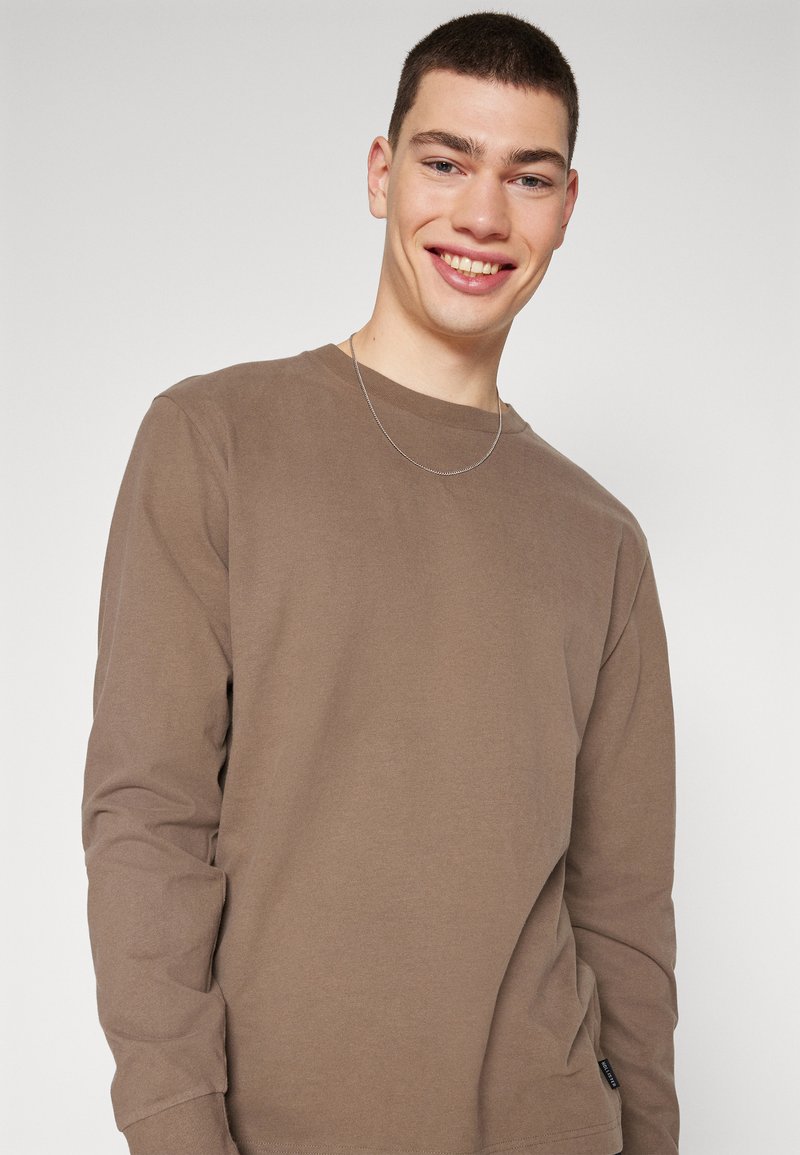 Braunes Langarm-T-Shirt aus weichem Stoff mit rundem Halsausschnitt und einem kleinen Markenlabel an der Seite. Einfaches, einfarbiges Design.