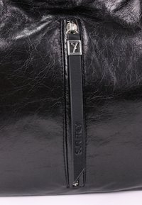 Gros plan d’un sac en cuir noir texturé avec une fermeture éclair verticale et une languette en cuir noir embossée "SURFREY."