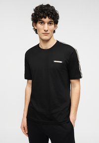 Μαύρο βαμβακερό T-shirt με κοντά μανίκια, στρογγυλή λαιμόκοψη και λογότυπο στο αριστερό μέρος του στήθους. Διαθέτει αντίθετες ρίγες στους ώμους.