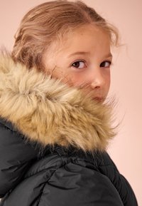 Černá péřová bunda s béžovým lemováním kapuce z umělé kožešiny. Má jemnou strukturu a přiléhavý střih. Pozadí je světle růžové.