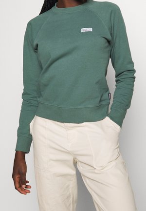 Persona que lleva una sudadera Patagonia verde bosque y pantalones de color crema, de pie con una mano en el bolsillo frente a un fondo liso.