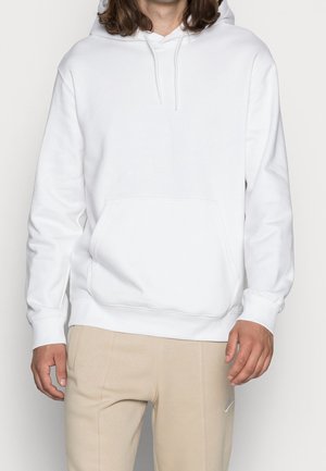 Person trägt einen schlichten weißen Hoodie mit Fronttasche und beige Hose, steht vor einem einfarbigen weißen Hintergrund.