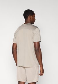 Αθλητική μπλούζα ανοιχτού μπεζ χρώματος με διχτυωτό κομμάτι στο επάνω μέρος της πλάτης, κοντά μανίκια και λεία υφή. Φοριέται με αντίστοιχα σορτς.