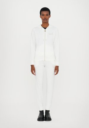 Tuta bianco a due pezzi con felpa con zip e cappuccio, tessuto morbido e dettagli con logo, abbinata a pantaloni affusolati e stivali neri.