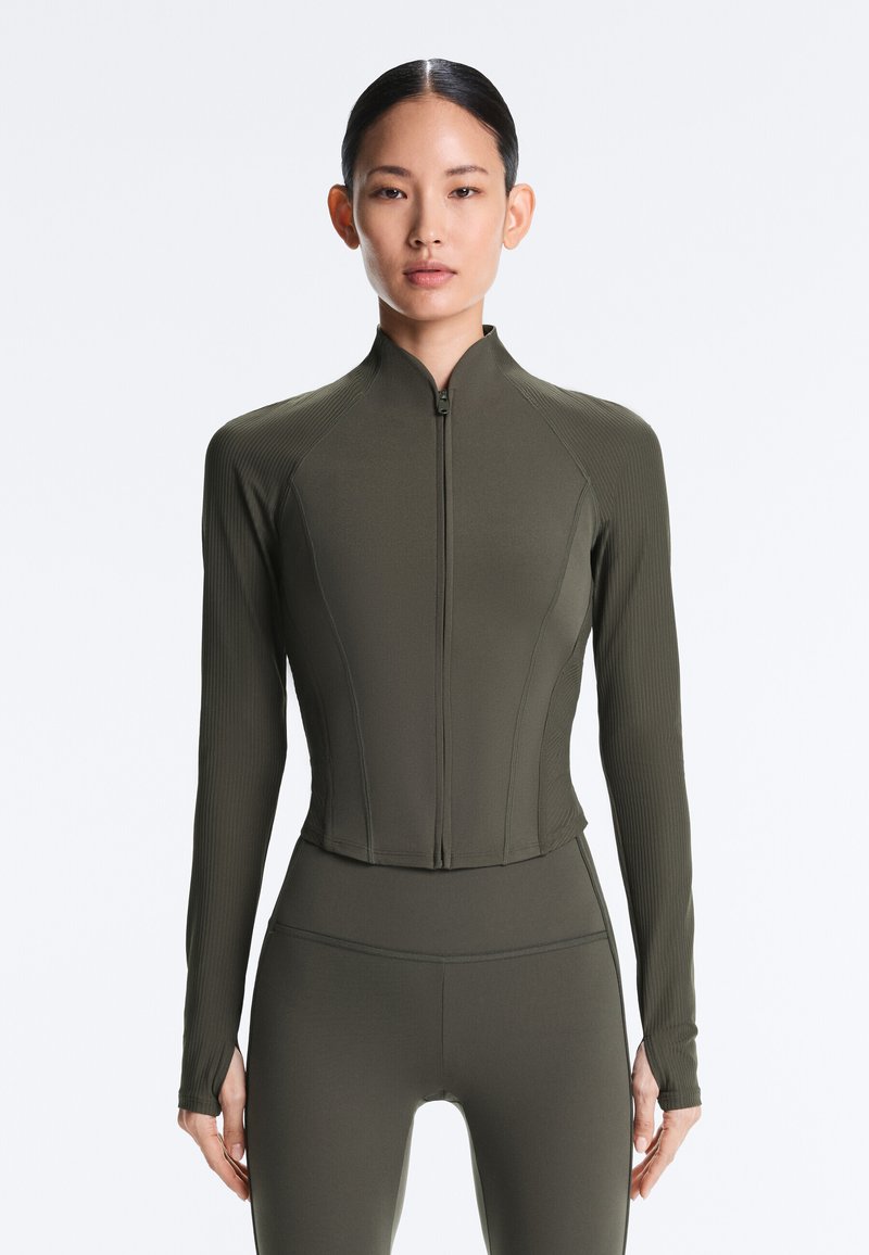 Femme portant une veste ajustée en olive à fermeture éclair avec manches longues et un legging assorti taille haute, debout devant un fond blanc.