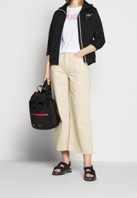 Chaqueta negra con cremallera sobre una camiseta blanca con texto rosa, pantalones de pierna ancha beige, mochila negra con detalle en cremallera roja y sandalias negras.