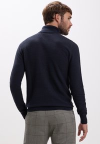Mann mit kurzen braunen Haaren, der einen strukturierten navyblauen Pullover und graue karierte Hosen trägt. Er steht mit dem Rücken zur Kamera und dreht den Kopf nach rechts.