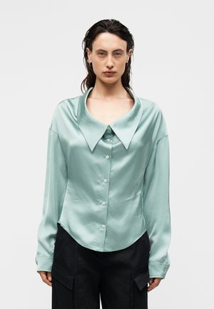 Frau trägt eine helltürkisfarbene Satinbluse mit weitem spitzem Kragen und Knöpfen, kombiniert mit schwarzen Hosen, vor einem weißen Hintergrund.