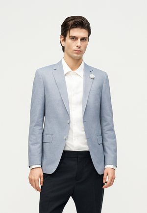 Junger Mann mit dunklen Haaren, der einen hellblauen Blazer, ein weißes Hemd und schwarze Hose trägt, steht vor einem schlichten hellen Hintergrund.