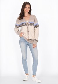 Beige kötött kardigán kék és barna mintázattal, V-nyakkivágással, gombos elülső résszel és két oldalsó zsebbel, világos farmernadrággal és fehér sportcipővel párosítva.