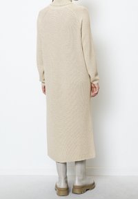 Femme portant un long manteau beige en maille et des bottes à semelle épaisse gris clair, debout contre un mur blanc.