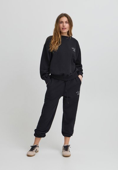 Completo felpa nera e joggers in morbido tessuto, con polsini a costine e un piccolo logo frontale. Il modello si presenta con le mani in tasca, indossando delle sneakers.