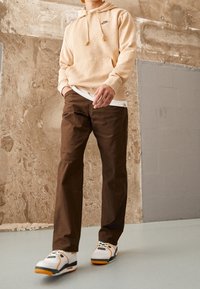 Beige hoodie with drawstrings and logo, layered over a white shirt; brown pants; white sneakers with orange and green accents.
