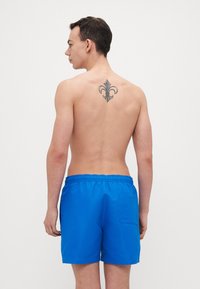 Blaue Badehosen mit einem elastischen Bund und einer Seitentasche. Das Modell ist oberkörperfrei und zeigt ein Tattoo auf dem Rücken.