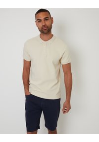 Camisa polo beige con un tejido texturizado, tapeta de cuatro botones y mangas cortas, combinada con pantalones cortos navy. Fondo liso.