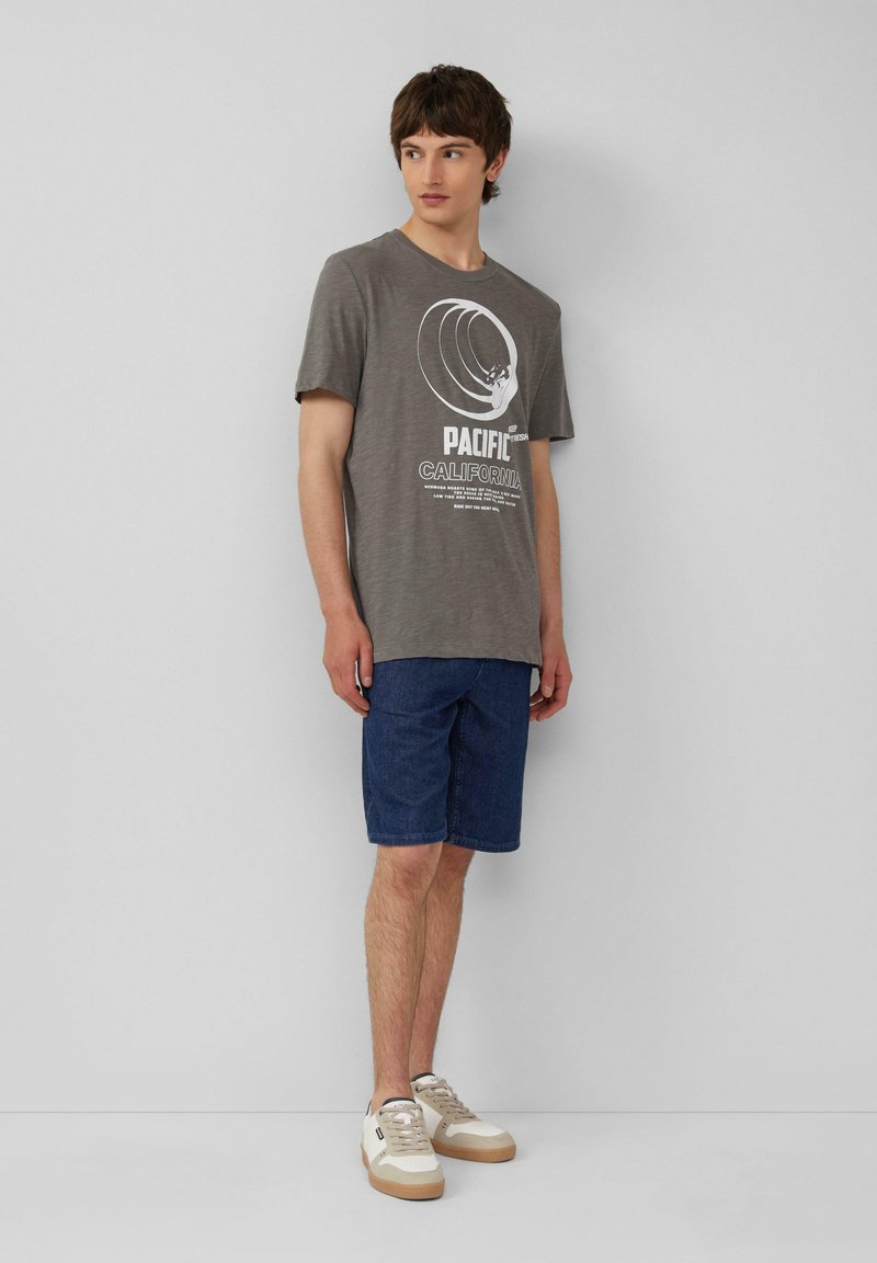 Grijze katoenen t-shirt met een witte grafische print, gecombineerd met donkere denim shorts. Beigekleurige sneakers met witte accenten maken de outfit compleet.