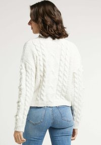 Pull en tricot blanc à motif texturé et accents bouffants, présentant une coupe courte et un ourlet côtelé, associé à un jean bleu.