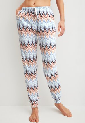 Vrouw in zachte, patroon joggingbroek met een chevronontwerp in blauw, bruin en zwart, staand op blote voeten tegen een effen achtergrond.