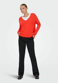 Femme portant un pull orange vif à col en V par-dessus une chemise blanche, un pantalon noir droit et des chaussures marron foncé, debout avec les mains dans les poches.