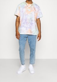 T-shirt w stylu tie-dye w pastelowych kolorach (fioletowy, pomarańczowy, miętowy) o luźnym kroju, z okrągłym dekoltem i haftowanym detalem. Połączony z jasnoniebieskimi dżinsami i białymi trampkami.