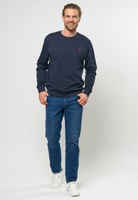 Marineblauwe sweatshirt met een ronde hals en ribgebreide manchetten, gecombineerd met een blauwe spijkerbroek en witte sneakers, staande op een lichte achtergrond.