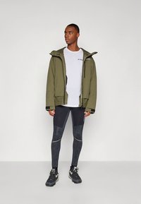 Vanngrønn vanntett jakke med hette, kombinert med en hvit t-skjorte og svarte mønstrede leggings. Sorte treningssko fullfører antrekket.