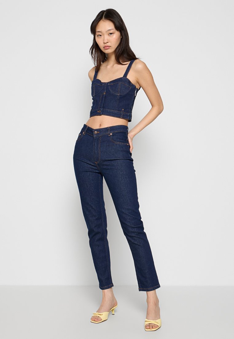 Jeansowa bluzka w kroju crop z szerokimi ramiączkami i guzikami z przodu, zestawiona z wysokim stanem, prostymi spodniami dżinsowymi w ciemnoniebieskim kolorze, dopełniona żółtymi sandałami na obcasie.