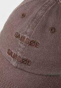 Cappellino da baseball in cotone marrone con logo ricamato "SAMSØE". Il tessuto ha una finitura testurizzata con cuciture visibili e fori di ventilazione.