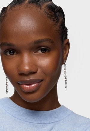 Zbliżenie na kobietę z plecionymi włosami, noszącą długie srebrne kolczyki w formie łańcuszka i jasnoniebieski sweter, delikatnie się uśmiechającą na białym tle.