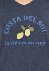 Granatowa koszulka z dekoltem w serek z żółtym graficznym motywem cytryny i zielonymi liśćmi. Niebieski tekst głosi "COSTA DEL SOL" oraz "la vida es un viaje."