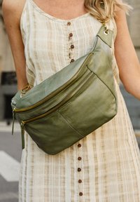 Borsa a tracolla verde in pelle con due scomparti zip, accessoriata con dettagli in oro e dalla texture liscia, indossata su un vestito a righe beige.