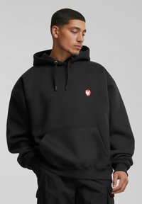 Schwarzer Hoodie aus dickem Stoff mit einer Vordertasche, verstellbaren Kordeln und einem kleinen roten Logo mit einem Apfeldesign auf der Brust.
