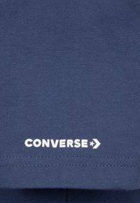 Sötétkék pamut póló fehér "CONVERSE" logóval az alján. Simább textúrával, látható minták vagy díszítések nélkül.