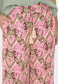 Pantalon ample à motif abstrait rose, vert olive et crème, taille à cordon avec pompons, mains partiellement dans les poches.