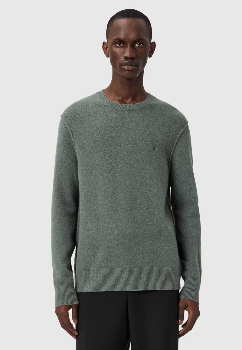 Mann trägt einen gedämpften grünen Pullover mit Rundhalsausschnitt und schwarze Hose, steht vor einem schlichten hellgrauen Hintergrund.