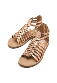 Sandalias gladiadoras en oro rosa con tiras trenzadas, suela plana y hebilla en el tobillo. Material texturizado y diseño abierto para una mejor transpirabilidad.