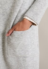 Hellgrauer Strickpullover mit geripptem Bündchen, Seitentasche und einem gestuften Look mit einem karierten Hemd darunter.